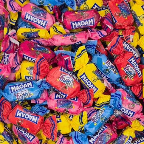 A clear plastic square container filled with assorted wrapped candies in bright pink, yellow, and blue packaging, photographed against a plain light gray wall.