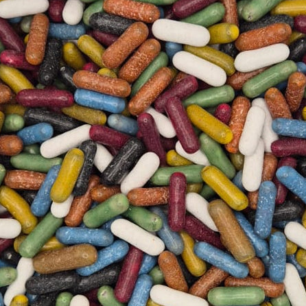 A clear plastic square container filled with many small multicolored capsules or pills, photographed from above on a white surface.