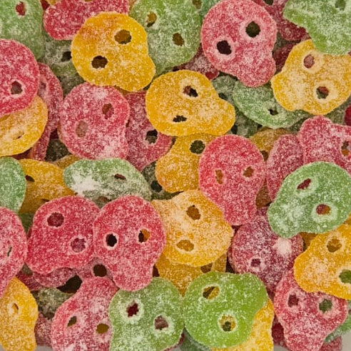 A clear plastic tub filled with small, sugar-coated, skull-shaped candies in pink, green, and yellow, photographed from above on a white surface.